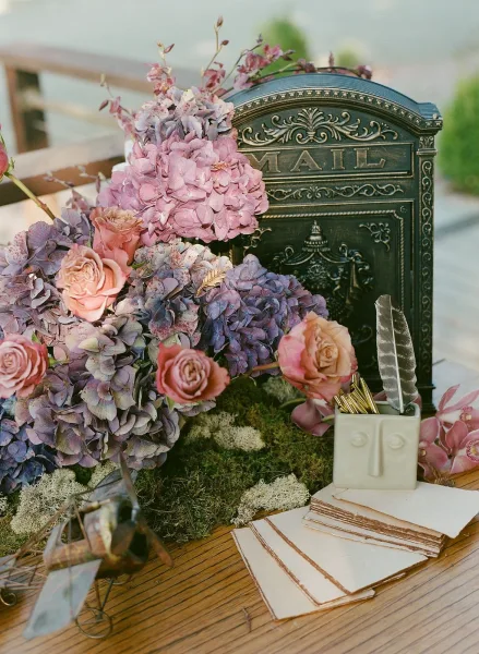 Wedding guestbook table with a wedding card box, ornate mailbox, hydrangeas and roses, wax seal stamps and envelopes on a wood table with greenery blurred behind