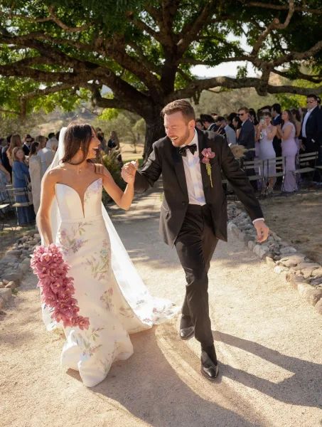 Wedding recessional as bride and groom walk down aisle holding hands, bride in strapless floral gown with veil, guests under trees
