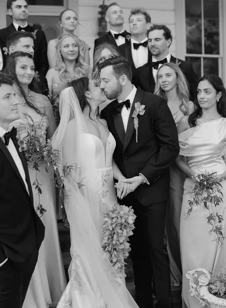 Wedding kiss portrait of the bride and groom kissing in black and white, her veil flowing as the wedding party stands on steps behind them