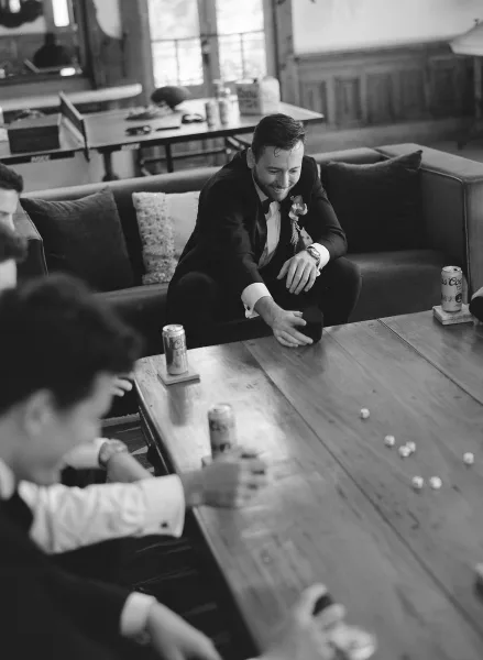 Groom getting ready in tuxedo laughs on a couch, boutonniere pinned, in a wood-paneled room with coffee table and windows