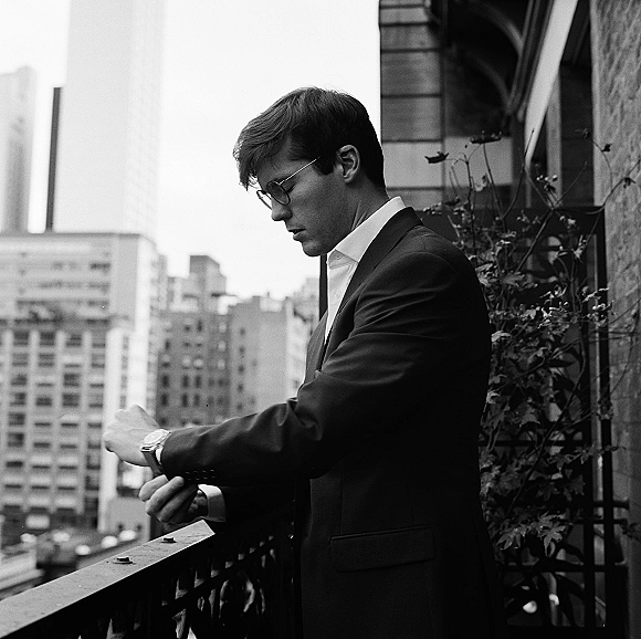 Groom portrait of a man in a suit adjusting cufflinks, wearing eyeglasses and wristwatch on a balcony with city buildings behind