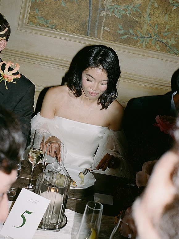 Reception moment of the bride at reception, seated in an off shoulder wedding dress at a table with glasses and a candle, mural wall behind