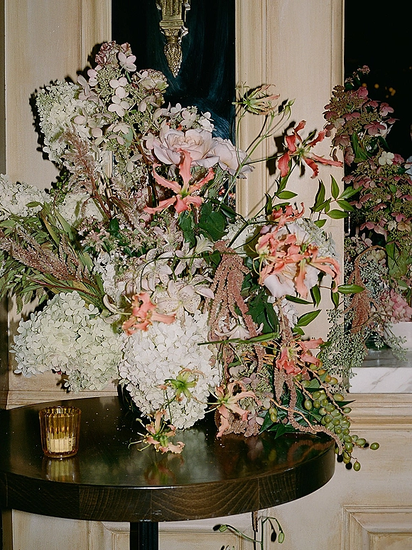Wedding floral arrangement with a large hydrangea centerpiece, pink and white roses and lilies with trailing greenery on a pedestal table, amber votive candle by a paneled wall mirror