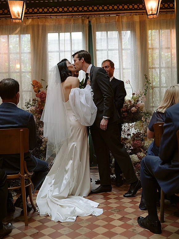 Ceremony kiss as bride and groom kiss beneath pendant lantern lights, her long veil and satin train flowing in window-lit indoor aisle