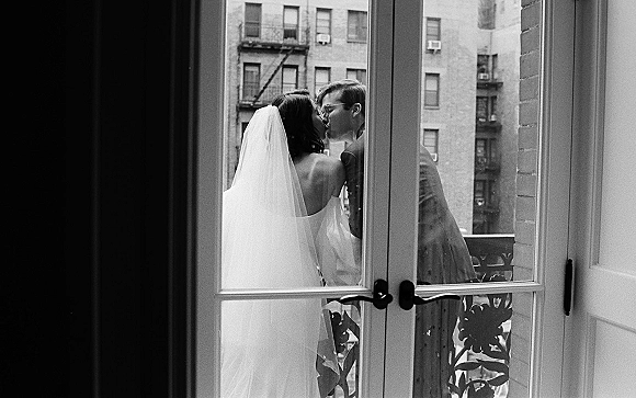 Wedding kiss portrait in black and white of bride in strapless dress and veil kissing groom in glasses on a balcony by French doors