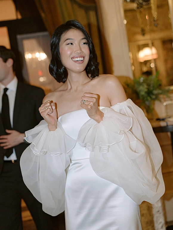 Bride portrait with a satin off-the-shoulder wedding dress and puff sleeves, showing rings under warm reception lighting with chandelier behind
