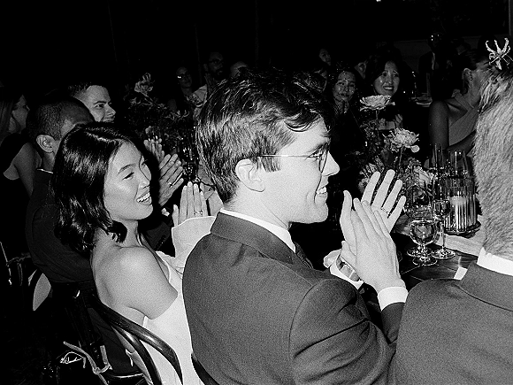 Wedding reception moment as guests applaud at a long table, champagne flutes raised amid floral centerpieces in dim indoor light
