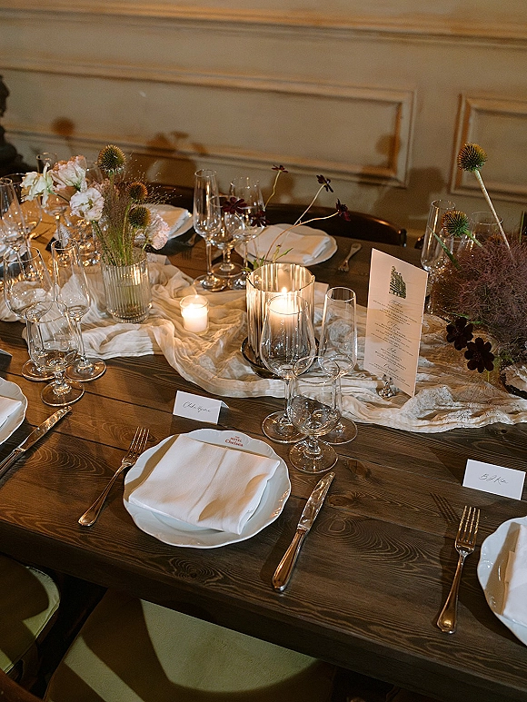 Reception tablescape with wedding table setting on a rustic wood table, white plates, silver flatware, stemware, candles, and floral vase against paneled wall