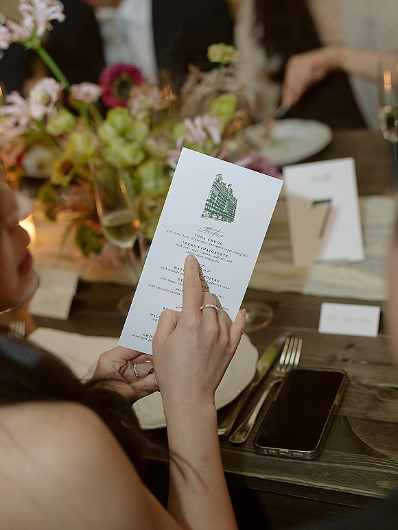 Wedding menu card held over a wood reception table with floral centerpiece, champagne flute, votive candle, and rings nearby