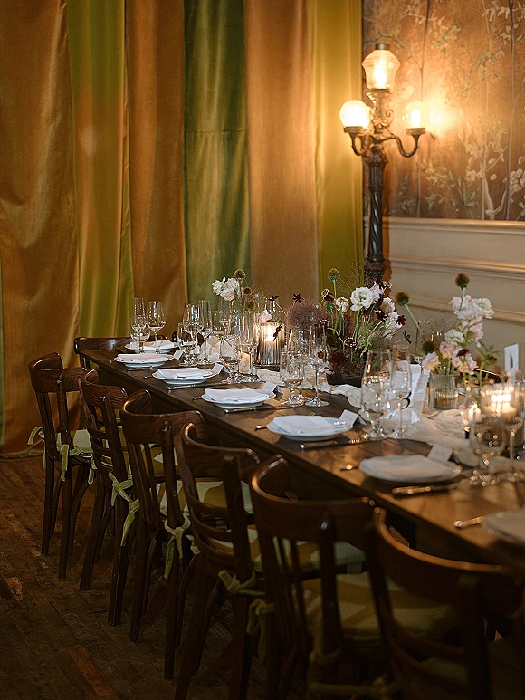 Reception tablescape on a long banquet table wedding setup with white plates, wine glasses, votive candles and low floral centerpieces in a draped room