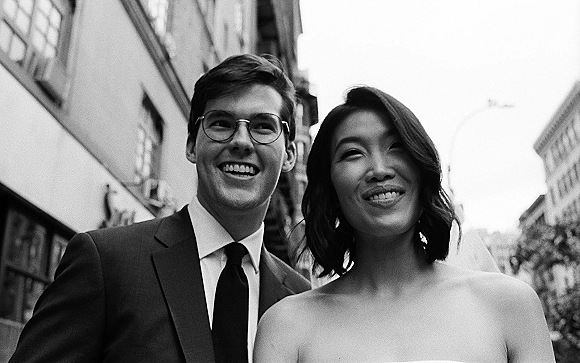 Couple portrait in a black and white wedding portrait style, bride in strapless dress and veil smiling beside groom in dark suit and glasses downtown