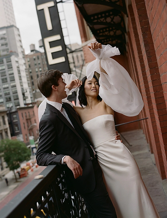 Couple portrait of bride in strapless satin wedding dress and groom in tuxedo leaning on balcony railing by a brick wall downtown