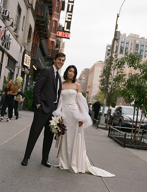 Couple portrait on a city street wedding photo, bride in strapless gown with puff sleeves holding bouquet beside groom in dark suit and glasses