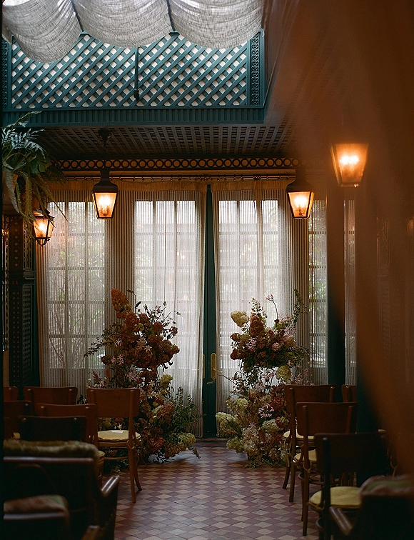 Ceremony setup for an indoor wedding ceremony with flower-lined aisle arrangements, chairs, and lantern pendant lights in a glass conservatory