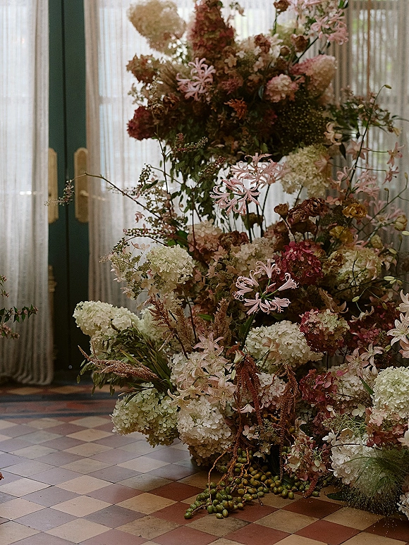 Wedding floral arrangement with hydrangeas and pink lilies in an asymmetrical design on a foam base by glass doors and sheer curtains