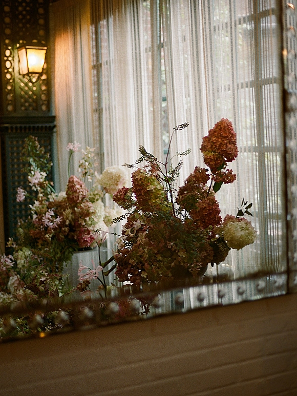 Wedding floral arrangement with hydrangea wedding centerpiece in a ceramic vase, arranged asymmetrically on a window ledge by sheer curtains