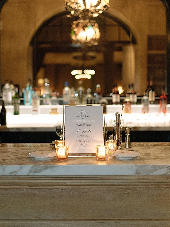 Wedding bar setup with a framed bar menu sign, votive candles, and glassware on the counter, backed by bottles under chandelier light