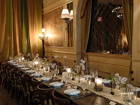 Reception tablescape with a long banquet table setup, taper and votive candles, bud vases, place cards and menus in an ornate room