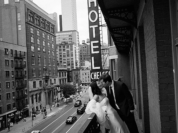 Wedding kiss portrait in a black and white wedding photo, bride in veil and strapless gown kissing groom by a balcony railing above a city street