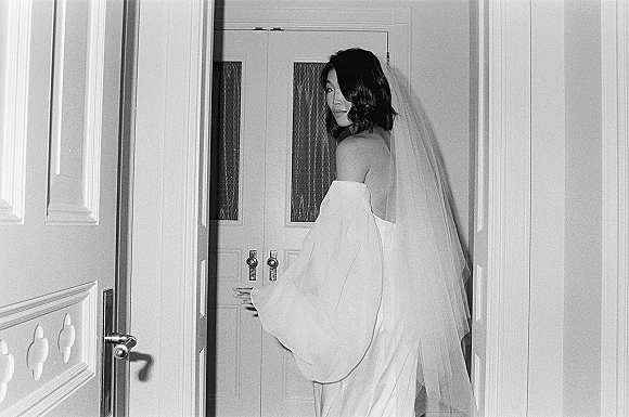 Bridal portrait in black and white of a bride looking back, wearing a strapless wedding dress and long veil by double doors in a hallway