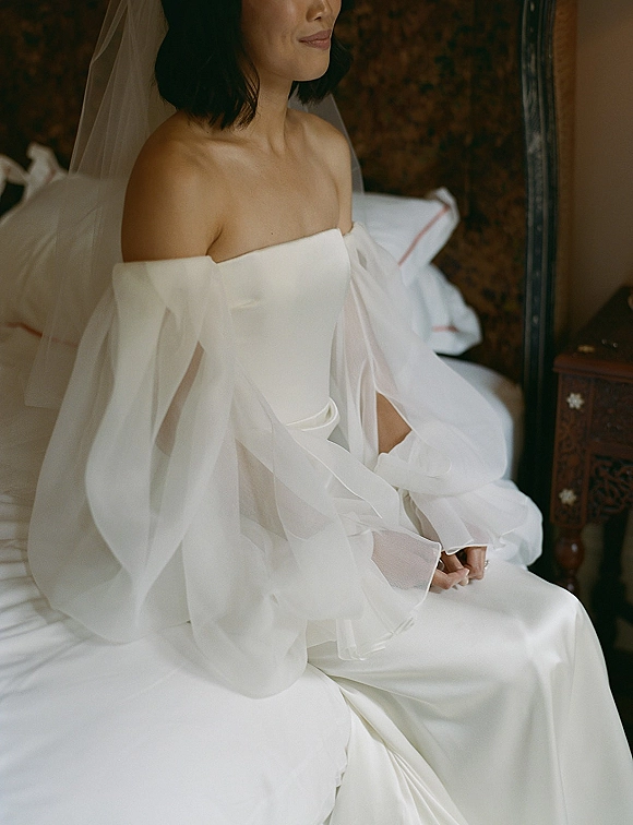 Bridal portrait of a bride sitting on bed in a strapless wedding dress with off-the-shoulder tulle sleeves and veil over shoulders