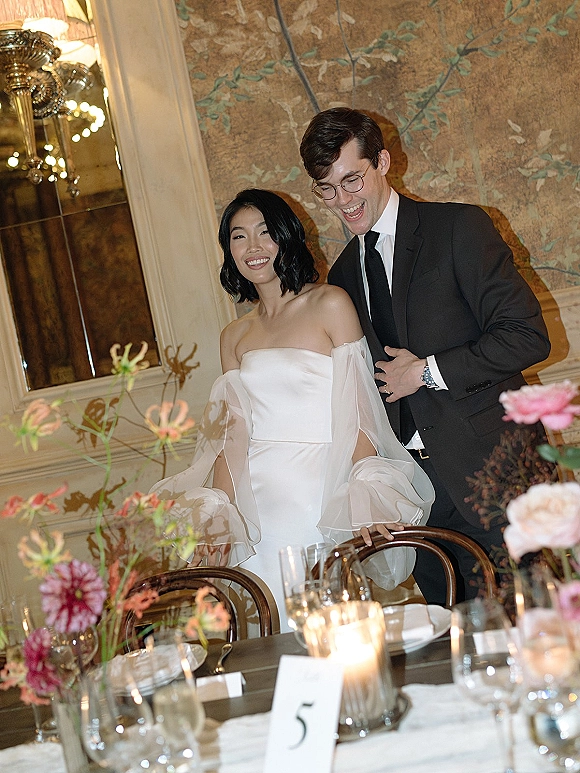 Couple portrait of bride and groom laughing as he hugs her by a candlelit reception table, with mural wall and floral centerpieces behind them