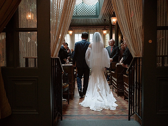 Wedding processional as bride walks down aisle with cathedral veil and long train, guests standing along checkered tile corridor under lantern lights