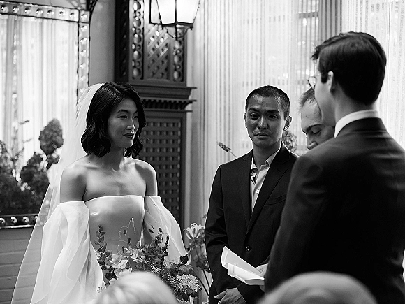 Wedding ceremony moment as bride in strapless dress and veil holds bouquet beside groom in dark suit during vows by large windows
