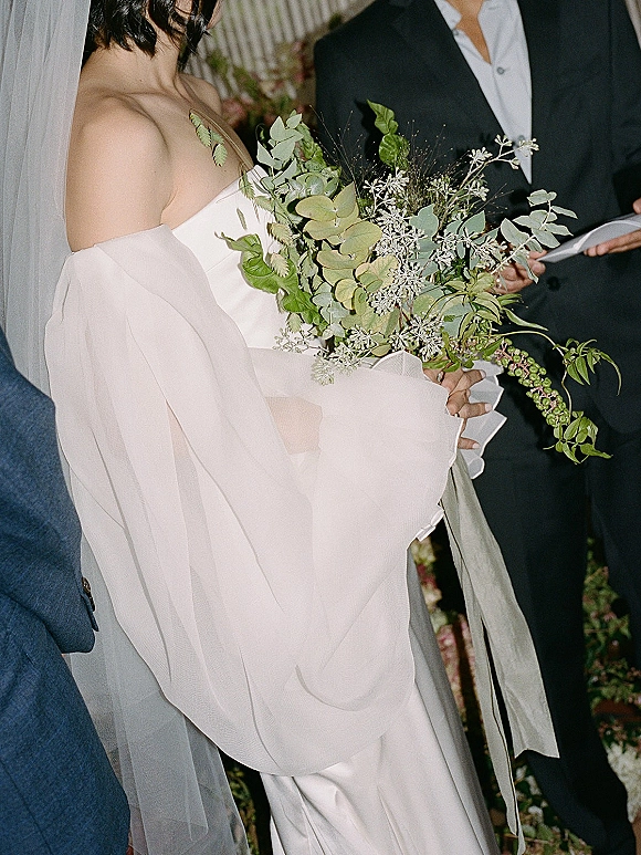 Ceremony moment as the bride holding bouquet stands in an off-the-shoulder gown with veil, beside groom in black suit indoors with florals