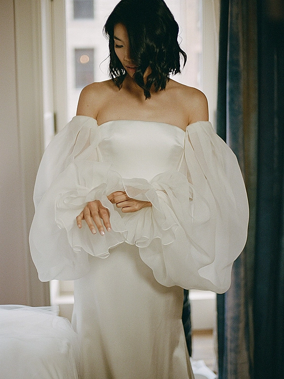Bridal portrait of a bride in an off the shoulder wedding dress with sheer organza puff sleeves, looking down by window light and curtains