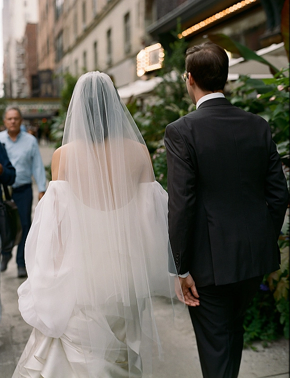 Wedding couple walking down a city sidewalk, bride’s long veil flowing behind her off-shoulder gown as groom in suit and glasses leads ahead