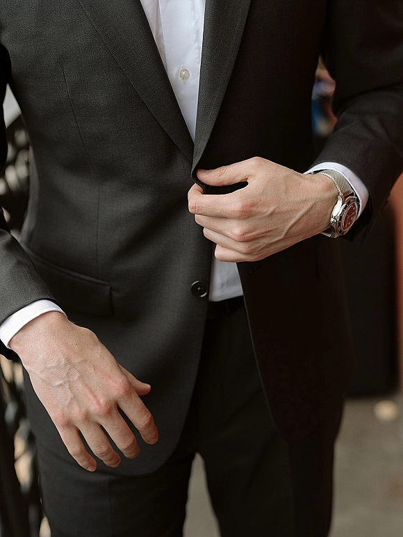 Groom suit details as he adjusts a dark suit jacket over a white dress shirt, showing a wristwatch, indoors near a railing