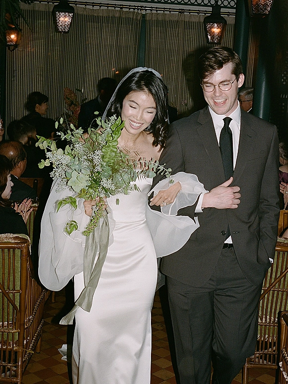 Wedding recessional as bride and groom walk up the aisle, bride holding a greenery bouquet with veil, guests in wooden chairs under lantern lights