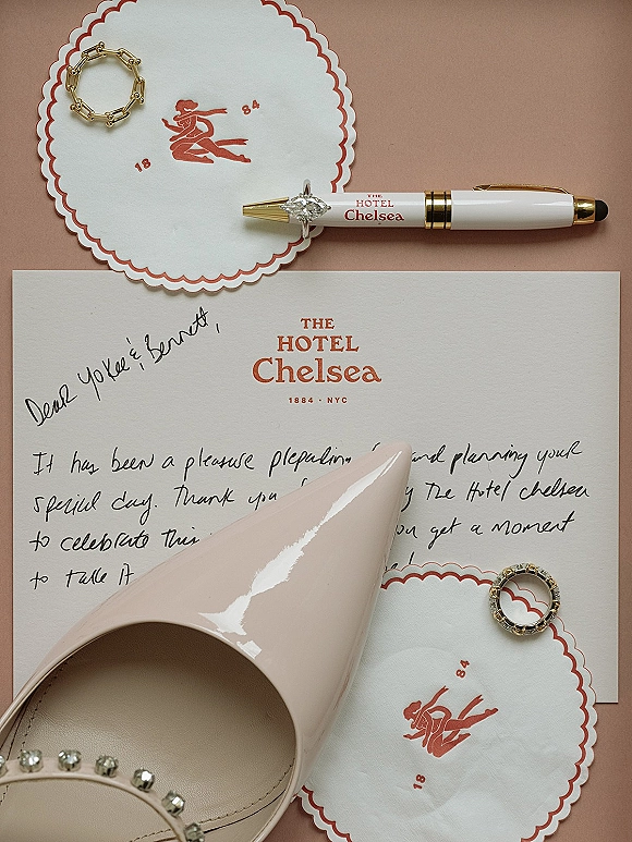 Wedding invitation suite with handwritten wedding note, rings, gold bracelet, scalloped napkins and bridal heel on a blush backdrop