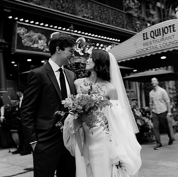 Couple portrait in a black and white wedding photo, bride with bouquet and veil gazing at groom in glasses on a city street by theater marquee