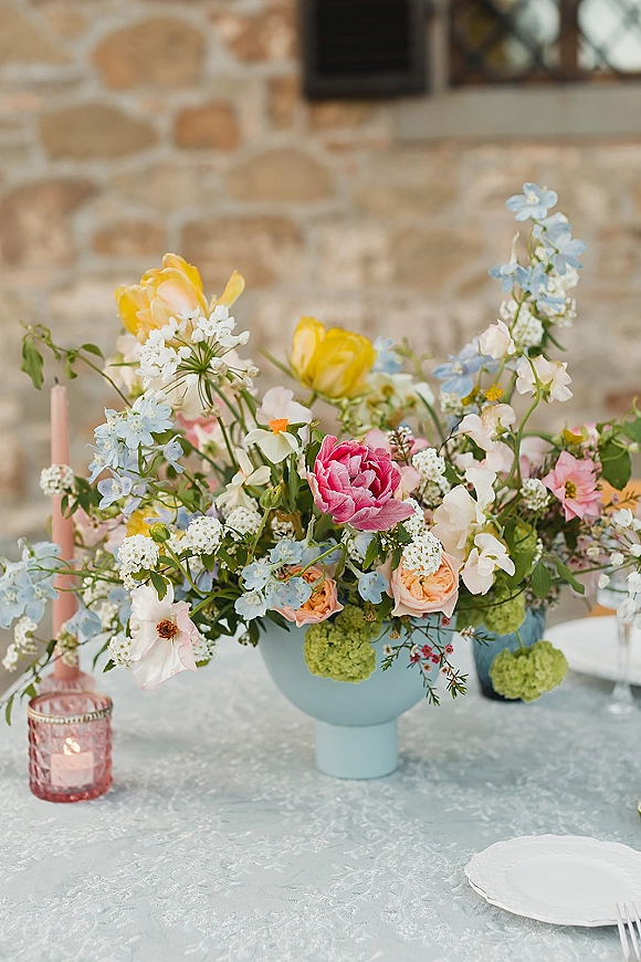 Wedding centerpiece with garden style flowers in a ceramic vase, pastel blooms and taper candle on a set table before a stone wall window