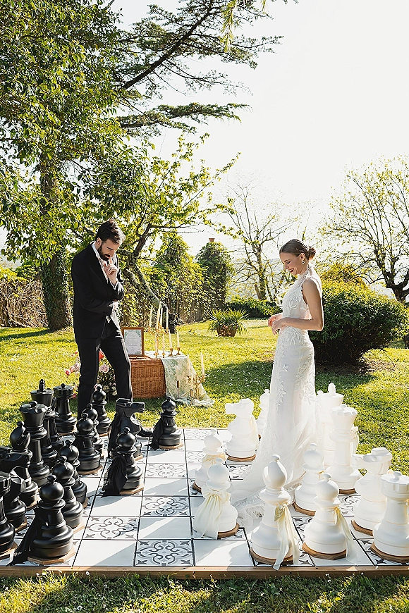 Couple portrait of bride in a high neck wedding dress and groom in tuxedo by an oversized chess set on sunny garden tiles, candles nearby