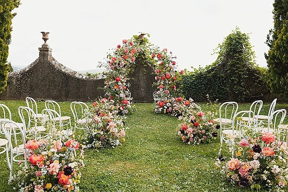 Ceremony setup with a floral ceremony arch, garden roses, and aisle flowers beside white bentwood chairs against an ivy stone wall