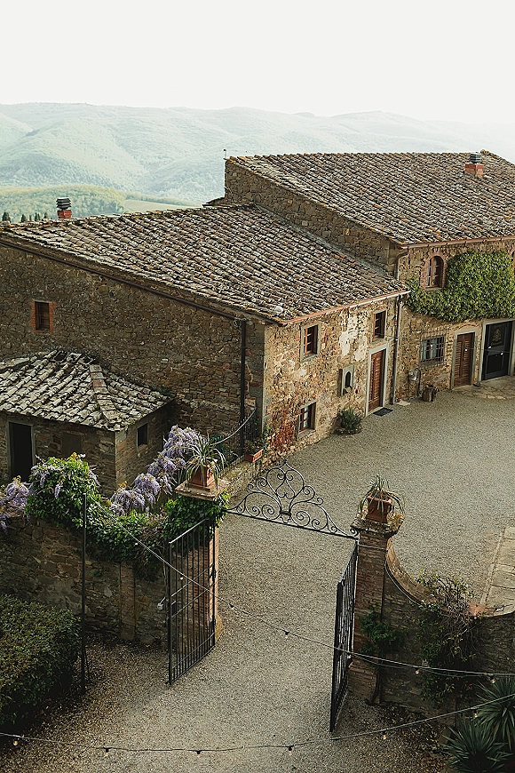 Wedding venue exterior of a stone villa with terracotta roof, wrought iron gate, gravel courtyard, and string lights against mountain hills