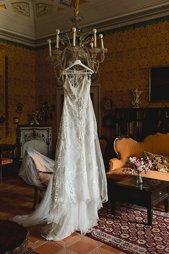 Wedding dress, lace wedding gown hanging from a chandelier in a vintage living room with fireplace, sofa, patterned wallpaper, and bouquet in vase
