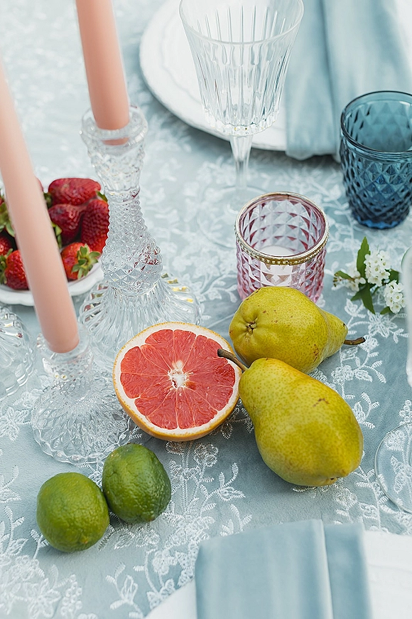 Reception tablescape with lace tablecloth, blush taper candles in cut glass holders, crystal stemware, fruit, and blue napkins on light blue linens