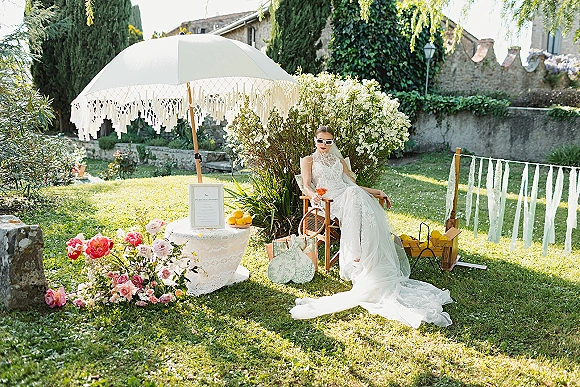 Bridal portrait of a bride in white sunglasses and lace high-neck wedding dress, seated with cathedral veil on a sunny garden lawn