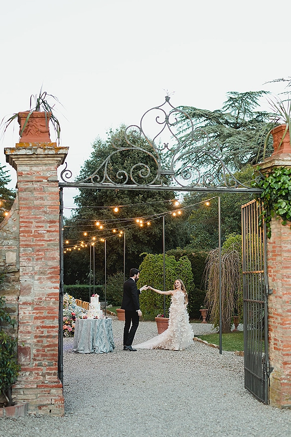 Couple portrait of bride and groom holding hands by a wrought iron gate with string lights, wedding cake table, and garden trees behind