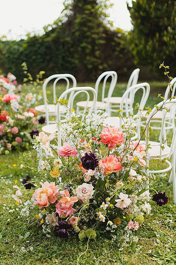 Ceremony aisle florals with grounded aisle flowers in coral and pink tones, lining white chairs on a grassy garden lawn with shrubs and trees