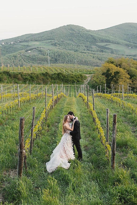 Couple portrait in a vineyard wedding photo, bride in strapless wedding dress and groom in tuxedo embracing among vineyard rows with mountains behind
