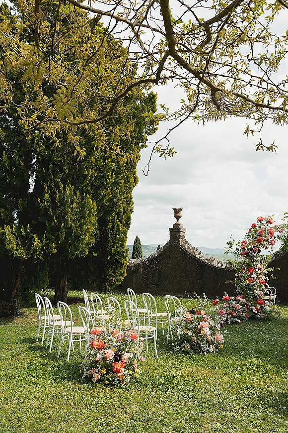 Outdoor ceremony setup with garden wedding ceremony seating, white chairs lining a flower-strewn aisle before an arch on a lawn by stone wall and trees
