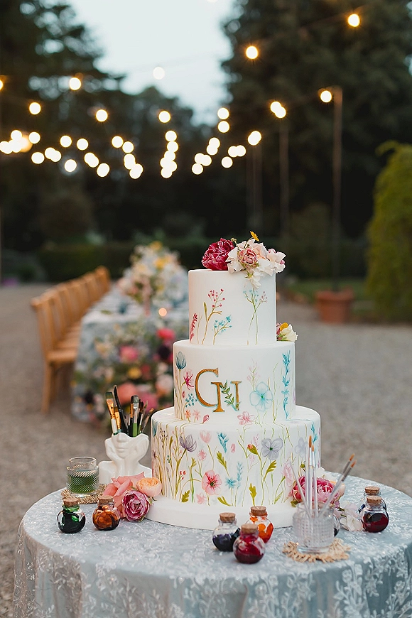 Wedding cake with hand painted floral design and monogram on a lace-covered table, fresh flowers and paintbrushes under string lights in a garden reception