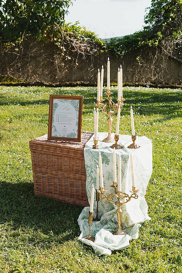 Wedding welcome table with a wedding welcome sign, gold candlesticks and white taper candles on patterned cloth beside a wicker trunk on a garden lawn