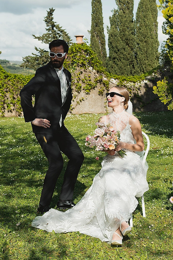 Couple portrait of bride and groom sunglasses, seated on a garden lawn by an ivy stone wall, bride in lace gown holding pastel bouquet
