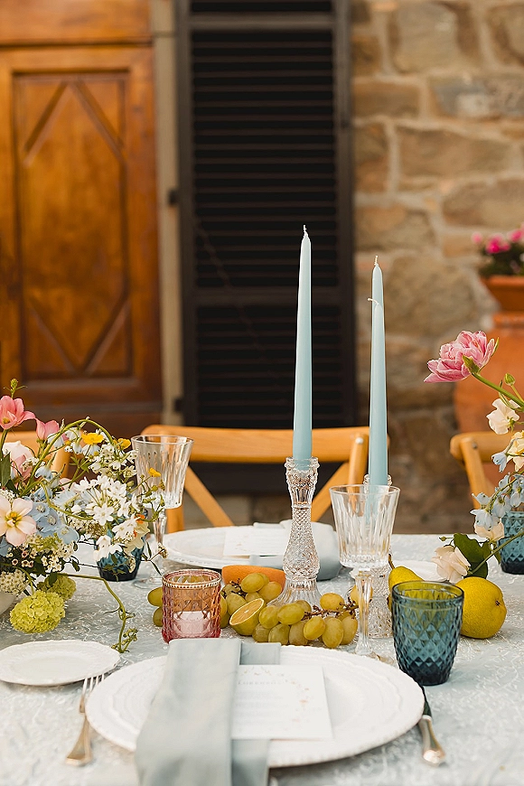 Reception tablescape with taper candles in crystal candlesticks, floral centerpieces, patterned linens, and fruit on a courtyard table by stone wall